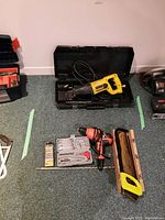 Dewalt reciprocating saw in open black case, Black & Decker drill, manual saw, and wrench set laid out on floor carpet