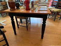 Side view of wood dining table with natural wood top and painted black legs and base. Shows scratches and wear on legs and floor beneath.