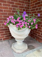 Front angle of a large cast polymer urn with light grey faux concrete finish, adorned with draped pattern, placed outside on stone floor against brick wall, filled with a live pink and purple flowering plant.