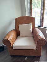 Front view of wicker armchair with beige cushions, showing woven arms and high backrest near a window with wooden blinds.