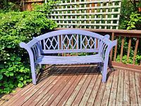 Full view of the light purple painted wooden bench on a wooden deck surrounded by greenery.