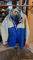Full view of the two-tone blue and beige Atlantis waterproof coat with hood hanging on wooden hanger.