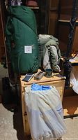 Photo showing green sleeping bag in compression sack, second sleeping bag rolled up, hatchet, green tin mug, Brunton compass, small boat anchor on wooden crate and inflatable air mattress bag in front.