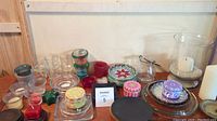 View of full lot showing multiple glass votive candleholders in various colors (clear, red, green), small decorative candles with patterned wrappers, and larger candleholders including a big hurricane glass and pillar candles.