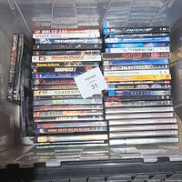 Top-down photo of DVD collection in clear plastic bin, titles mostly action and adventure films and some documentaries.