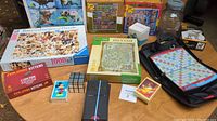 Full view of the lot with games and puzzles displayed on a table showing Exploding Kittens game, travel Scrabble, puzzles, and coin jar.