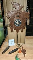 Front view of the cuckoo clock on a cardboard box showing carved bird, leaf motif, black clock face with Roman numerals, tangled chains, and pinecone weights on a wooden surface.