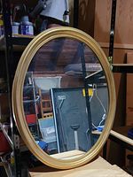 Gold oval wall mirror with gold frame leaning on shelving, showing the shape and frame style.