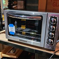 Front angled view of the KitchenAid stainless steel toaster oven on a metal shelf with a blue MaxSold lot label.