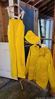 Full view of yellow waterproof overalls on hanger and yellow raincoat on hanger next to yellow rain hat on separate hanger, showing front details
