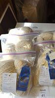 Five sealed Ziploc bags filled with off-white wool yarn skeins, including skeins with attached blue prize ribbons and handwritten tags
