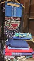 Full view of stacked textiles including tablecloths, runners, napkins, and towels in various colors and patterns such as blue, red, fruit prints, and holiday themes.