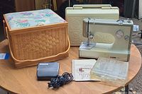Photo showing vintage Kenmore sewing machine, hard carrying case, woven sewing basket with cushioned lid, instruction manual, foot pedal, and other small items on a wooden table.