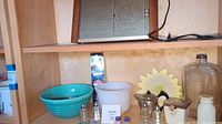 Shelf displaying turquoise Bauer 12 mixing bowl, white Glasbake mixing bowl, ribbed glass wine bottle, honey storage jar with sunflower lid, silver teapot, salt and pepper shakers from West Germany.