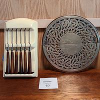 Photo of 6 steak knives with brown Bakelite handles in original cream stand next to round ornate silver plated trivet fully visible