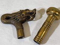 Two brass cane handle tops; one dragon shaped with detailed scales and mane, the other a traditional ball design with ring accents on shaft.
