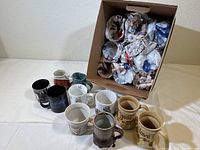 19 assorted coffee mugs shown partially outside of a cardboard box with additional mugs packed inside with newspaper wrapping.