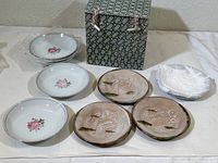Overview of both sets of small plates and bowls displayed outside of the cloth storage box, showing white floral plates on the left, brown ceramic plates on the right, and the green patterned cloth box in the background.