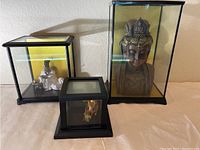Three figurines displayed inside black-framed glass display cases on a surface against a wall.