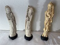 Three carved wood statuettes on black pedestal bases displayed against a white wall.