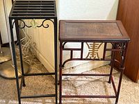 Two Far East style display tables: one faux bamboo wood table with woven top and Chinese character decoration, one black metal table with slatted top and decorative gold leaf front.