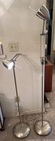 Two metal adjustable floor lamps standing side by side, showing height difference and metal base and heads.