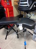 Two black adjustable music seats: one rectangular, one saddle-style, both on metal bases near a red cabinet and black chair