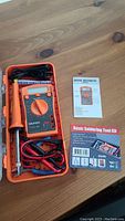 Open orange plastic case containing the Gold Tool GSM-881 digital multimeter with probes, soldering iron component and other accessories in foam insert, sitting on a wooden surface. Basic Soldering Tool Kit package and instruction manual beside it.
