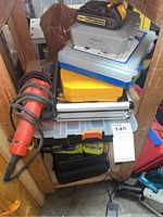Stack of tool cases and chargers on shelving, includes DeWalt charger and battery, Ryobi charger, Kreg jig case, yellow DeWalt bit case, red corded tool, Makita cordless drill visible