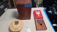 Shows two kalimbas and a wooden percussion instrument on a table, highlighting the rectangular wooden kalimba with red packaging and the circular percussion instrument with kalimba tines.