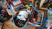 Box with assorted power tools including drills and a white handheld tool, cardboard boxes visible, various cords and hand tools mixed in.