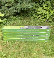 Four green labeled Gutterware Aluminum Gutter Protection System boxes stacked horizontally outdoors on grass with leafy plants in background.
