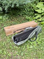 Black tarp rolled inside green carrying bag alongside a rectangular box labeled WAKEMAN Outdoors containing ground stakes.