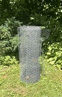 Front view of rolled chicken wire spool standing on grass with garden foliage in background, showing hexagonal wire mesh pattern.