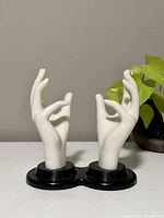 Two white plastic hands with fingers posed elegantly on connected black bases, shown against a gray background with a green plant.