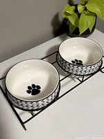 Two white stoneware dog bowls with black paw prints inside, sitting side by side in black metal stand from a front angle, showing full inside of bowls and design.