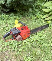 Red gas powered small chainsaw placed on grass with surrounding green plants. Chain has been removed from the cutting bar for safety.