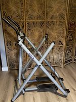 Front angled view of the silver Gazelle exercise machine showing foldable frame, handlebars, and foot pedals.