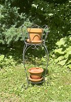 Full view of metal two-tier plant stand outdoors on grass with two terra cotta pots placed on each tier.