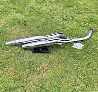 Pair of chrome motorcycle tailpipes laid on grass, showing full length and mounting bracket.
