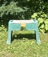 Front view of green plastic garden kneeler and bench with yellow clasp on hinged lid, photographed outdoors on grass with shrubs in background.
