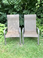Two taupe mesh sling patio chairs on green grass with leafy background, showing front view of chairs with armrests