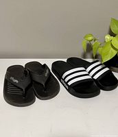 Two pairs of sandals side by side on white surface: Adidas black slides with white stripes and black Ginga flip-flops.