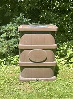 Front view of a brown rectangular rain barrel with Green Port logo outdoors on grass surrounded by plants.