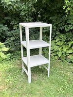 Full view of the three tier metal shelving unit standing outdoors in grass with bushes in the background.