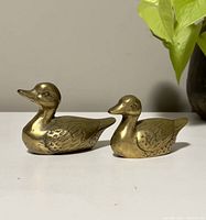 Side view of the two brass duck figurines, showing the larger duck on the left and smaller on the right against a neutral background with plant leaves above.