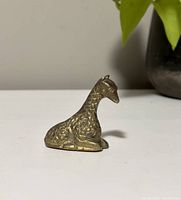 Side view of the brass giraffe figurine showing its detailed textured body and sitting posture on white surface with plant in background.
