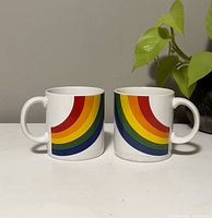 Pair of white ceramic mugs shown side by side, featuring a rainbow design with six color arcs.