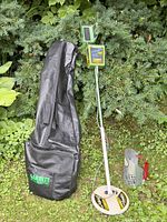 Full view of metal detector and accessories with carrying case on grass and pine shrub background