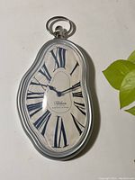 Front view of the melting clock showing distorted oval shape, Roman numeral hour markers, blue clock hands, and silver-tone frame with hanging ring on top.